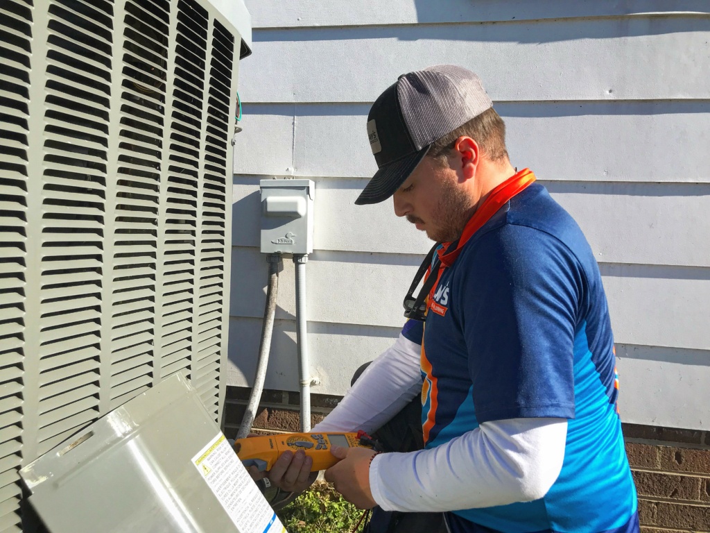A man in a blue shirt and cap works on an outdoor air conditioning unit using a handheld tool.