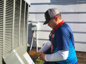 A man in a blue shirt and cap works on an outdoor air conditioning unit using a handheld tool.