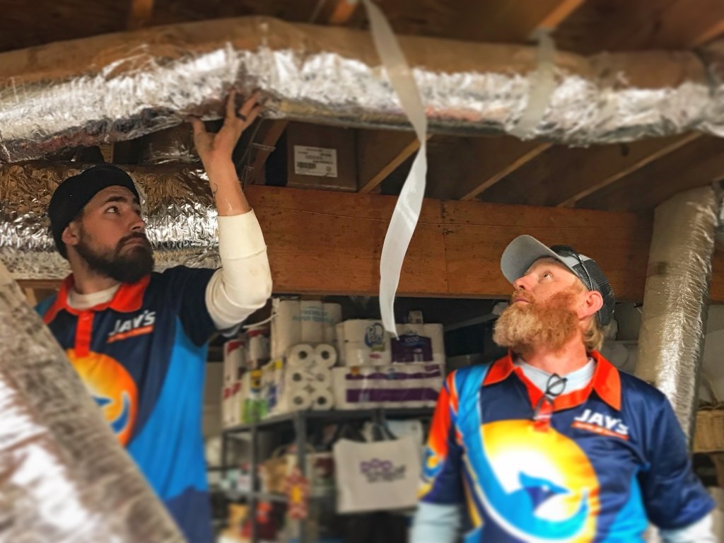 Technicians assess and work on ductwork in a basement or garage, surrounded by household supplies.