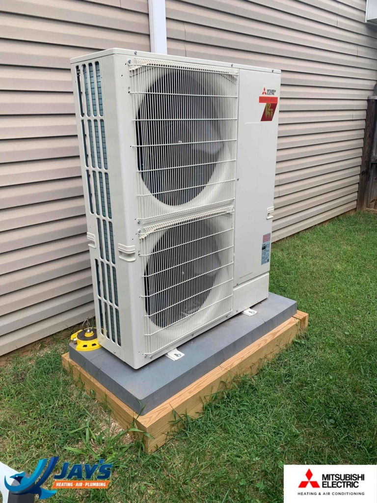 An outdoor air conditioning unit is mounted on a wooden platform next to the exterior of a house. The unit has two large fans covered by metal grilles.