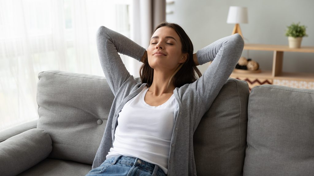A woman sits on a grey couch with her hands behind her head, eyes closed, appearing relaxed.