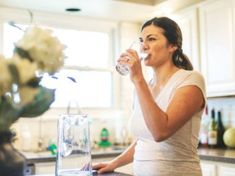 A woman in a light-colored shirt drinks water from a glass in a sunlit kitchen. A vase with flowers is on the countertop.