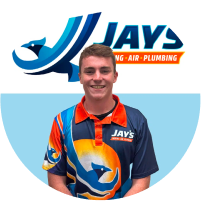 A man in a multicolored service uniform with the logo "Jay's Heating Air Plumbing" standing in front of a circular team logo backdrop.