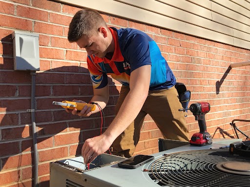 Technician checking an air conditioning unit with a multimeter outside a brick building on a sunny day.