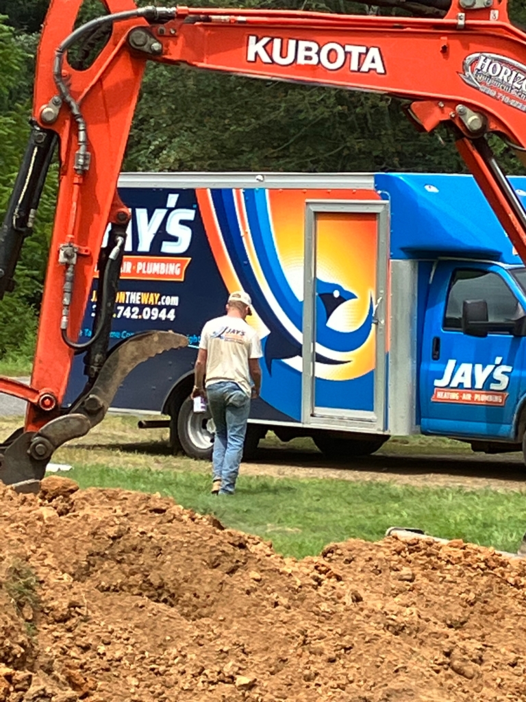 A person wearing a white shirt walks beside a bright blue and orange work truck labeled "Jay's," with an excavator arm visible in the foreground. Dirt piles are in the foreground.